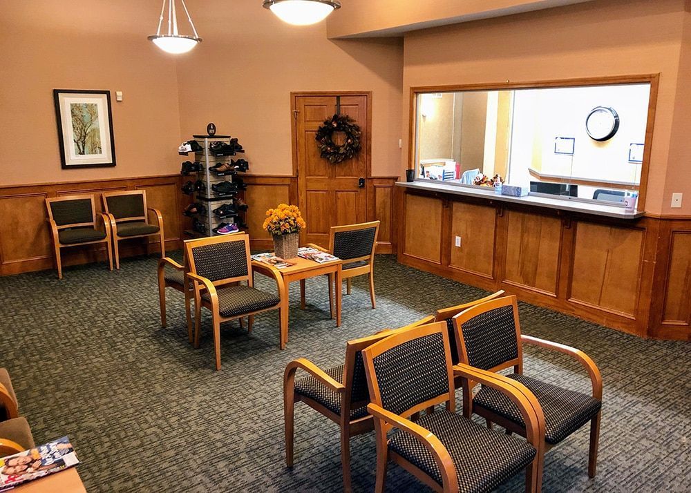 Waiting room with chairs, small table, and reception desk. Brown paneling and walls, neutral carpet.