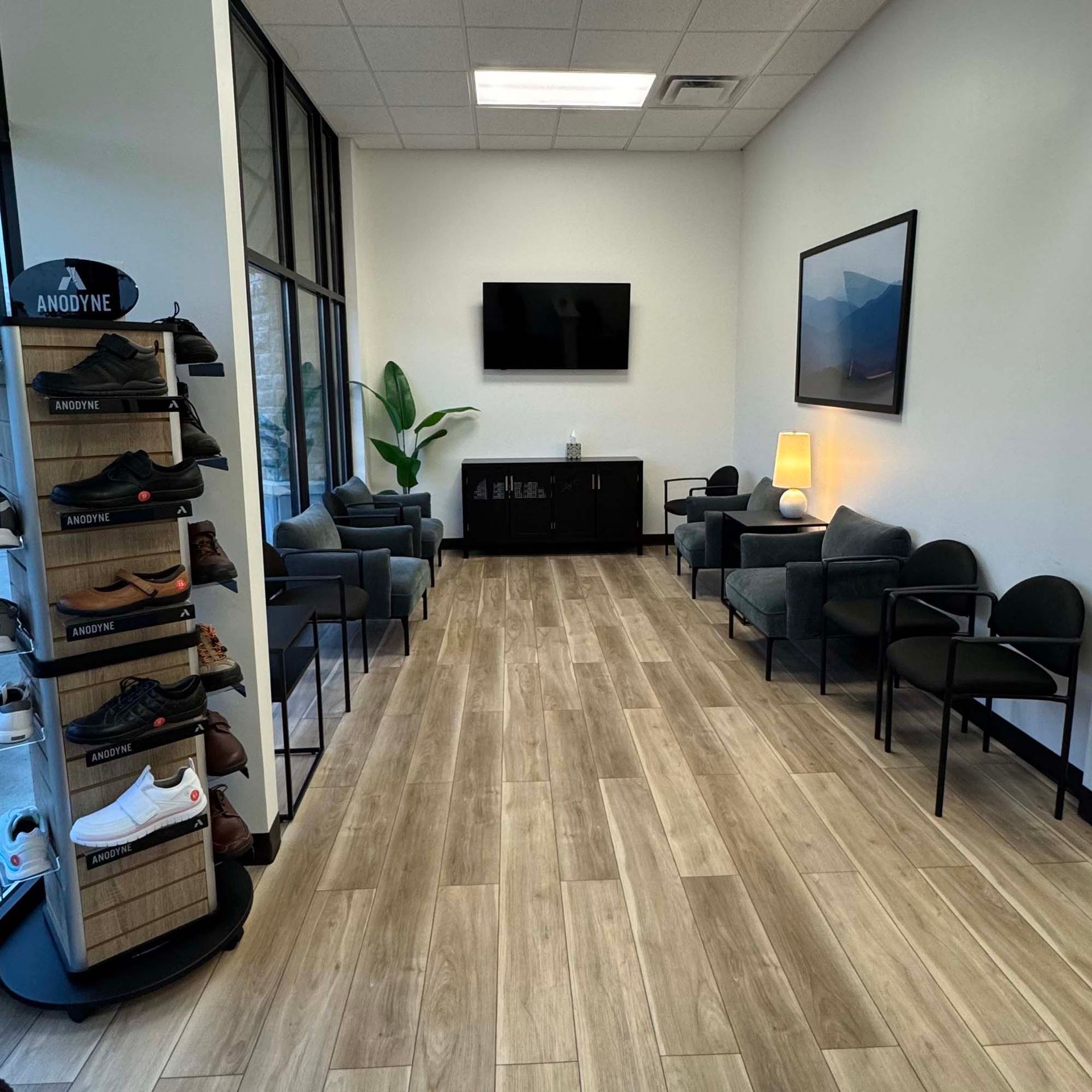 Waiting room with chairs, TV, and shoe display; light wood-look floor, white walls.
