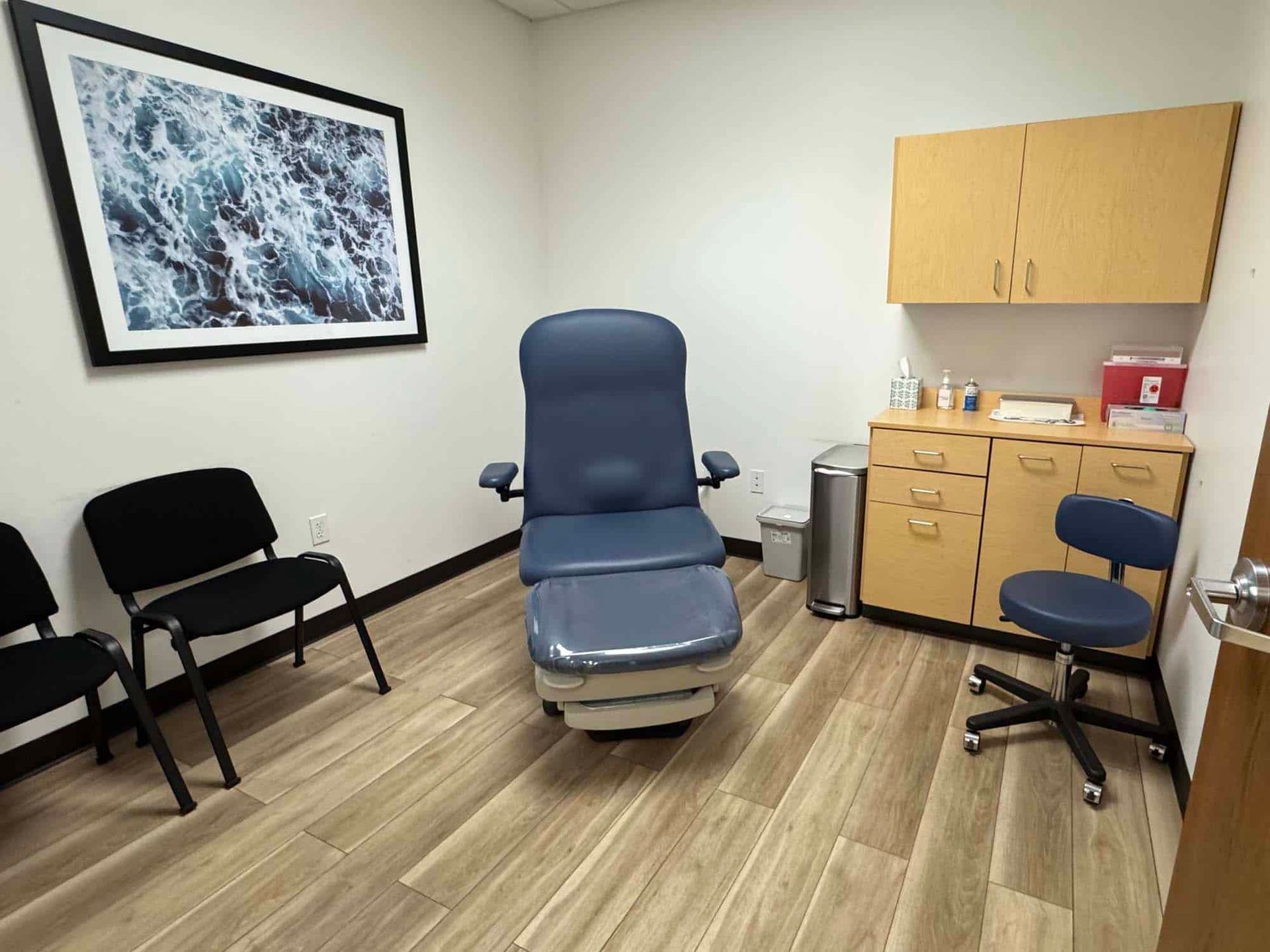 Medical exam room with examination chair, two black chairs, and cabinets. Artwork on the wall.