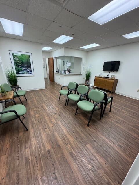 Waiting room with green chairs, wood-look floor, framed art, and a small TV. Reception desk visible.