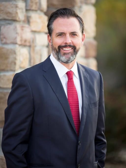 Man in a suit, red tie, smiling; standing by a stone wall outdoors.
