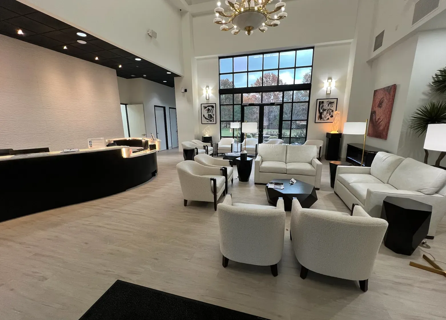 Lobby with white sofas, chairs, black tables, and a reception desk. A large window overlooks outdoor greenery.