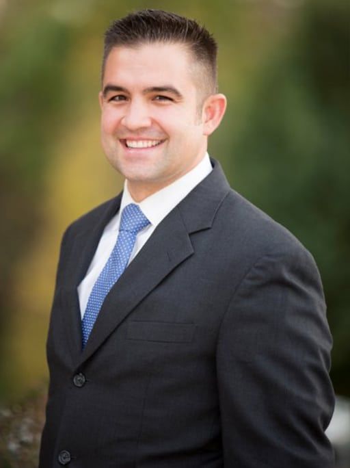 Man in a suit smiles, blue tie, short dark hair, outdoors with blurred greenery.
