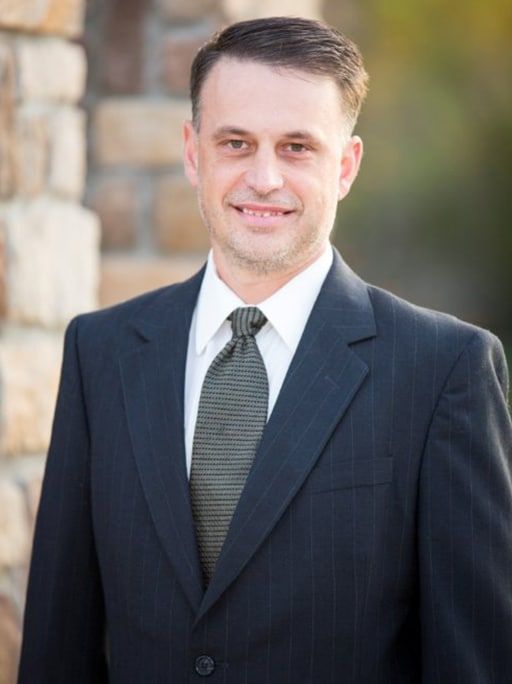 Man in a suit smiles outdoors, near a stone wall.