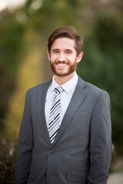 Man in gray suit and tie smiles against a blurred outdoor background.