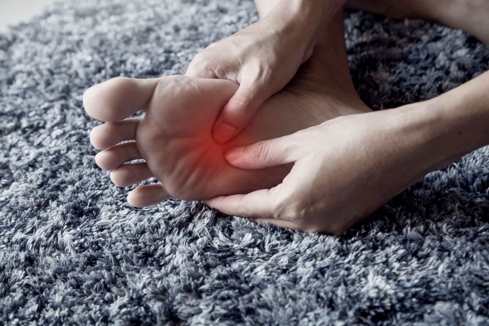 Person holding their foot, highlighting a red spot on the sole, possibly indicating pain on a gray carpet.