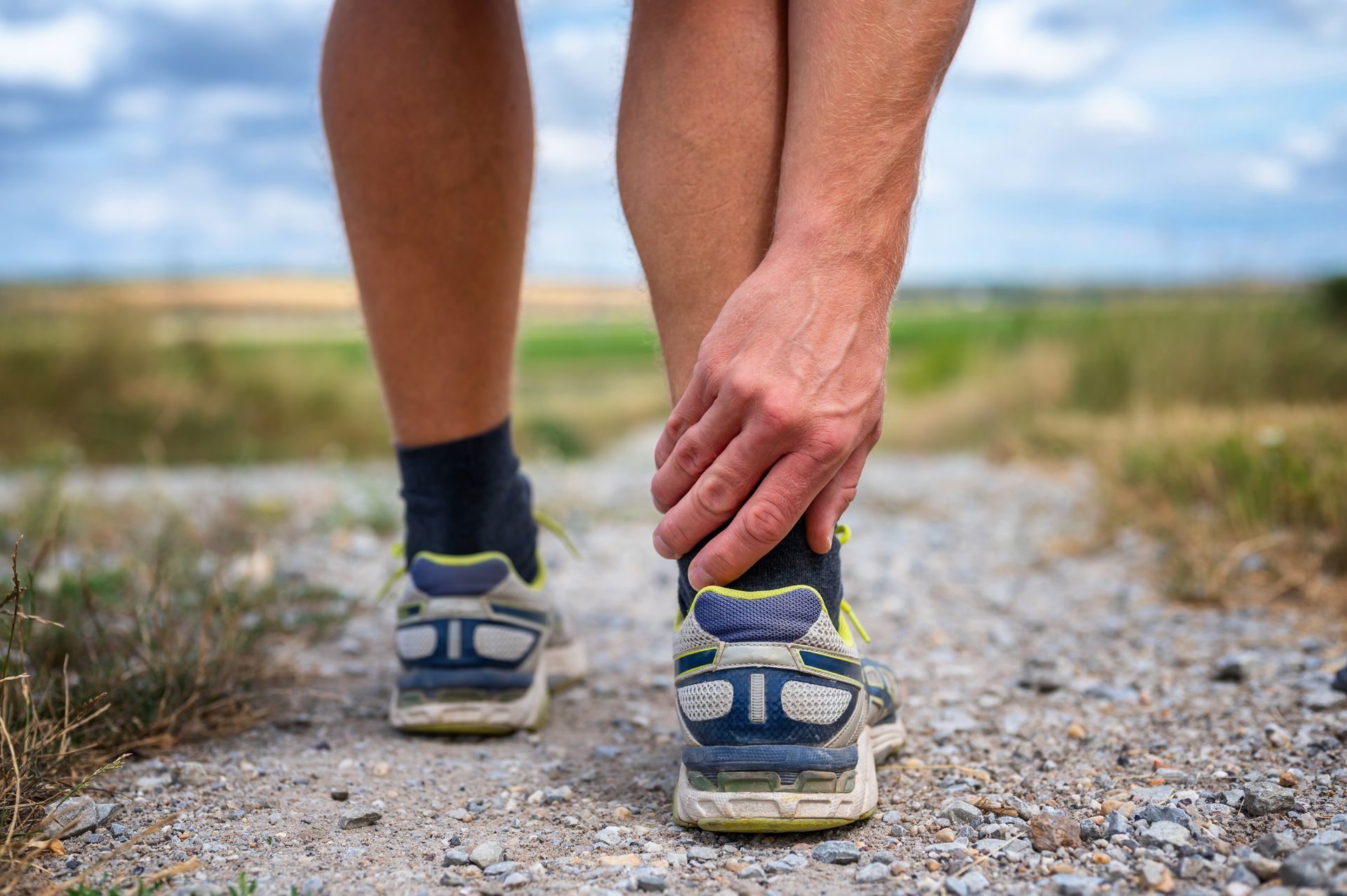Person clutching ankle while wearing running shoes on a dirt path.