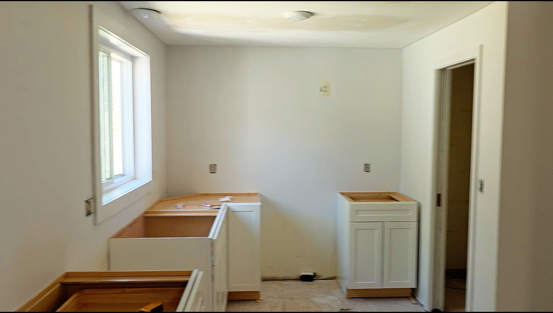 A kitchen under construction with white walls, window, and unfinished white base cabinets installed along the walls.