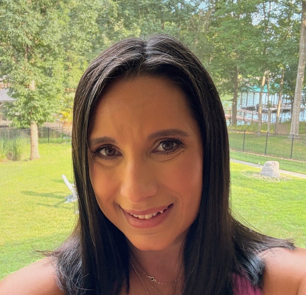 A person with dark hair smiling at the camera, set against a blurred background of a grassy yard with trees and a lake.