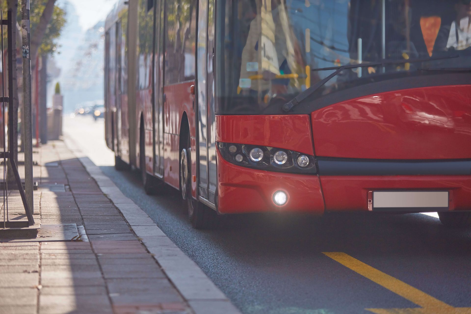 View of a large red city bus driving along a sunlit urban street with buildings in the background