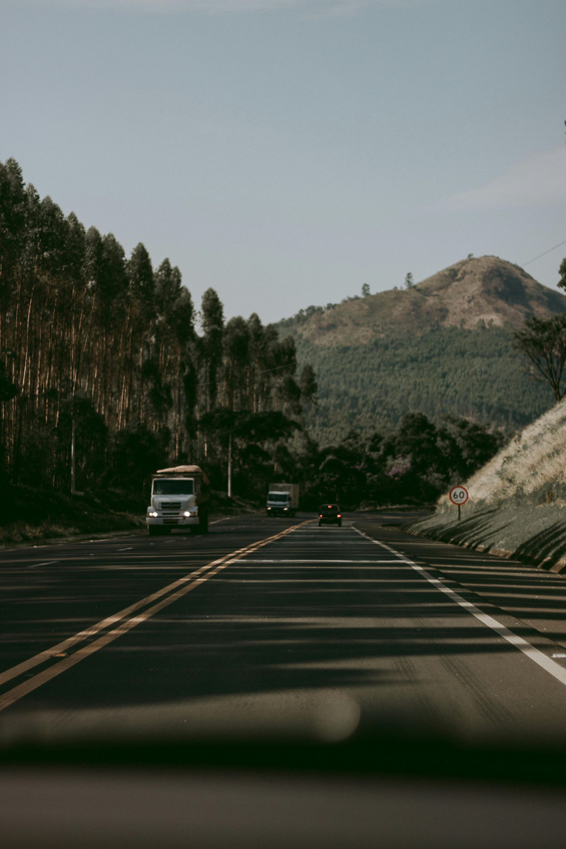 A scenic road with trucks and cars traveling through a mountainous landscape, lined with tall trees on one side and a hill in the background.