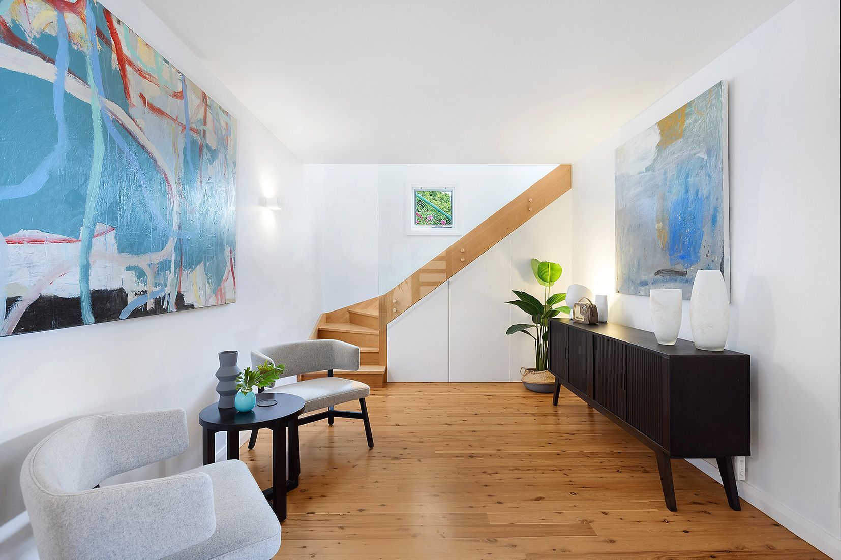 Bright hallway with artwork, wood staircase, and dark credenza. Light wood floors and two cozy chairs.