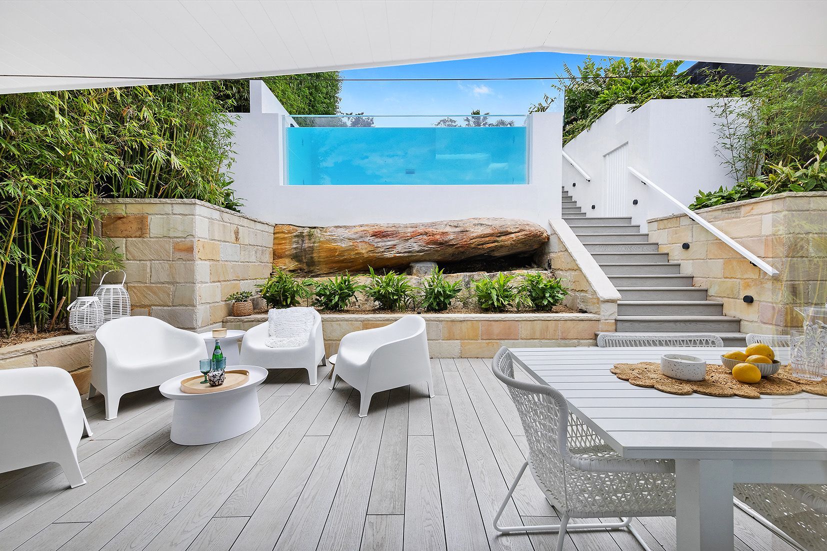 Patio with pool. White furniture on a wooden deck. Pool with clear side and stairs leading up.