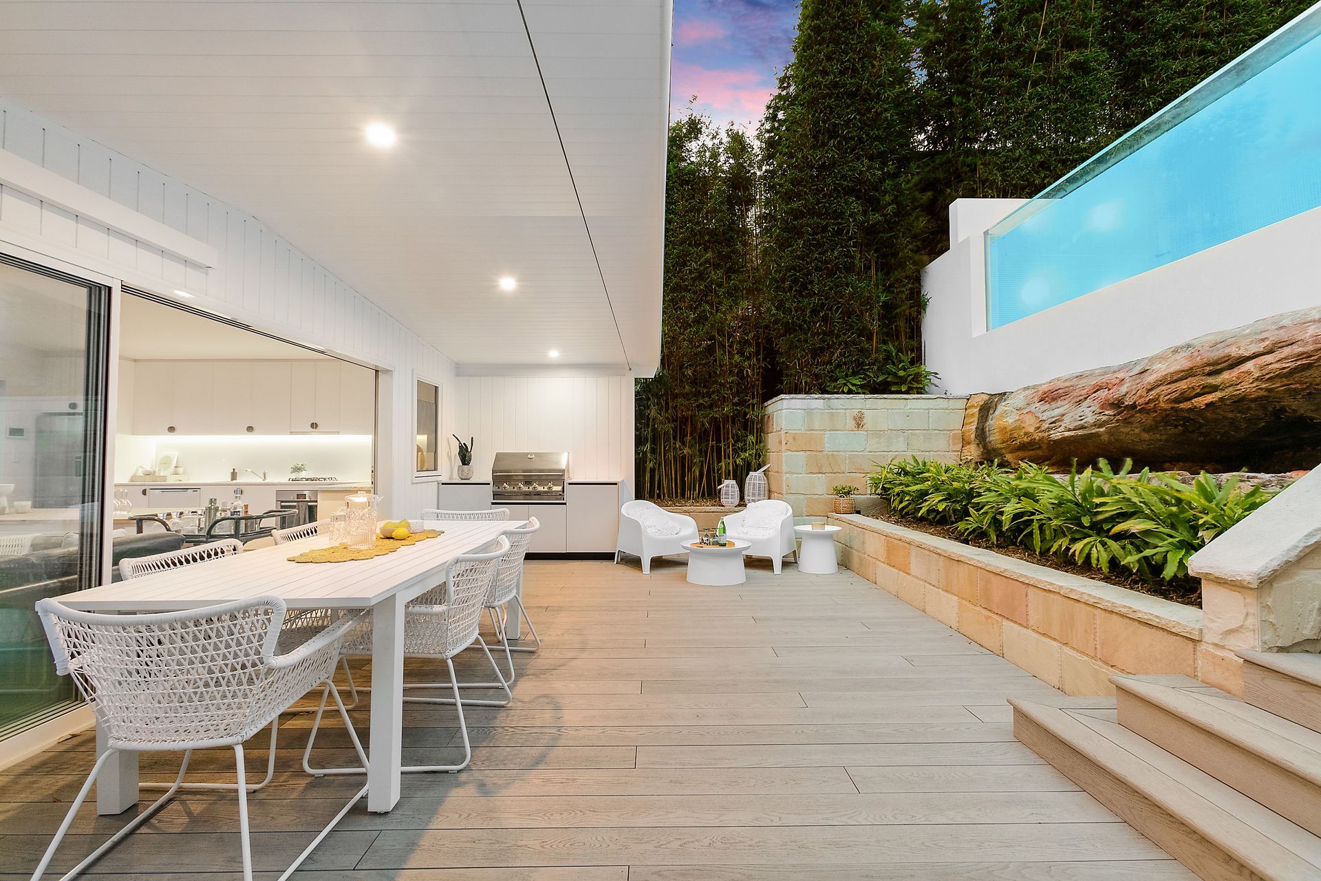 Outdoor dining area with white furniture, kitchen, and pool overlooking greenery.