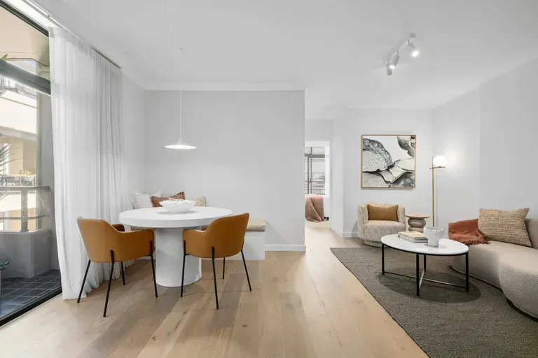 Brightly lit modern apartment with dining and living areas. Light wood floors, white walls, and orange chairs.