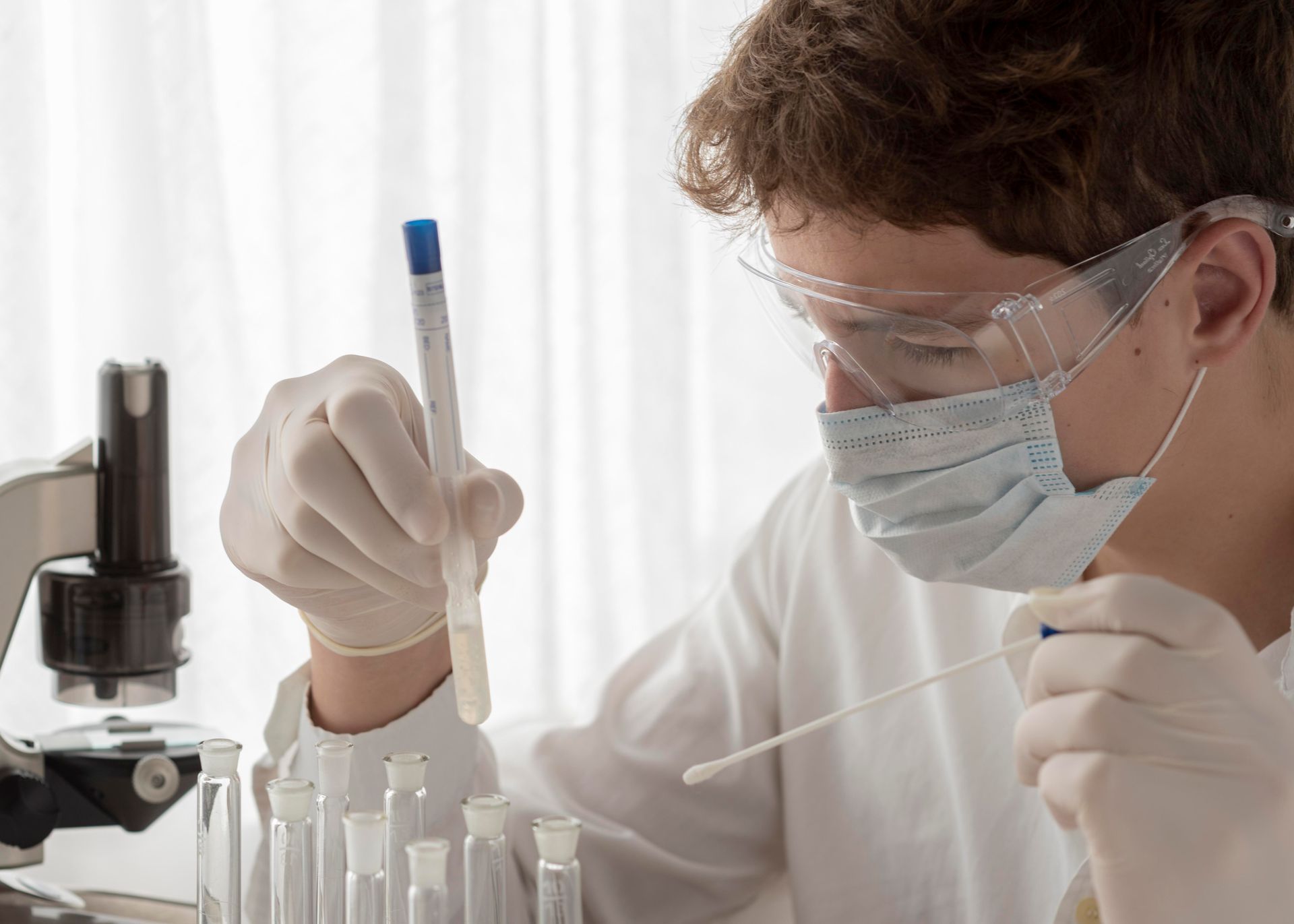 A man wearing a mask and goggles is looking at a test tube in a laboratory.