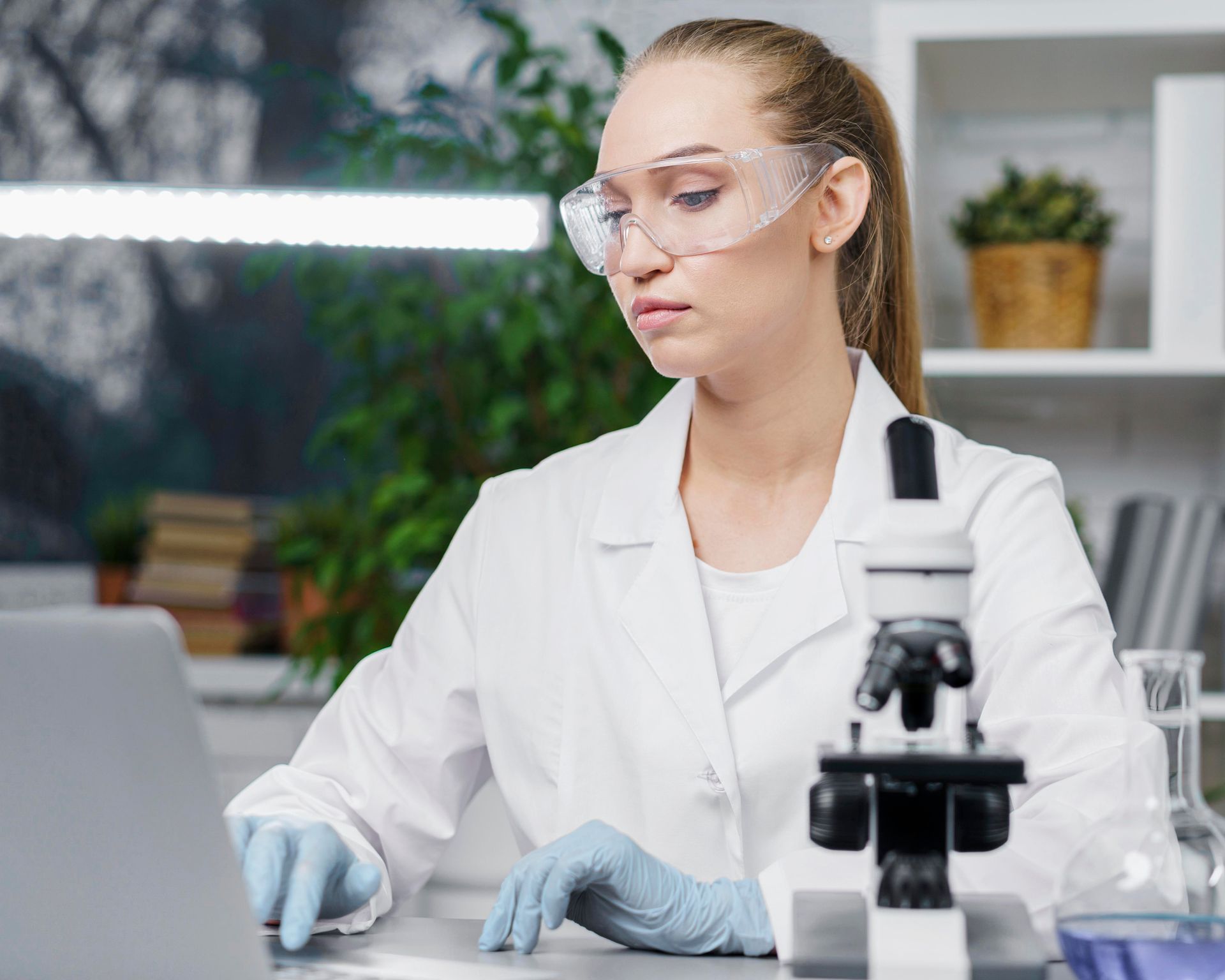 A female scientist is using a laptop and a microscope in a lab.