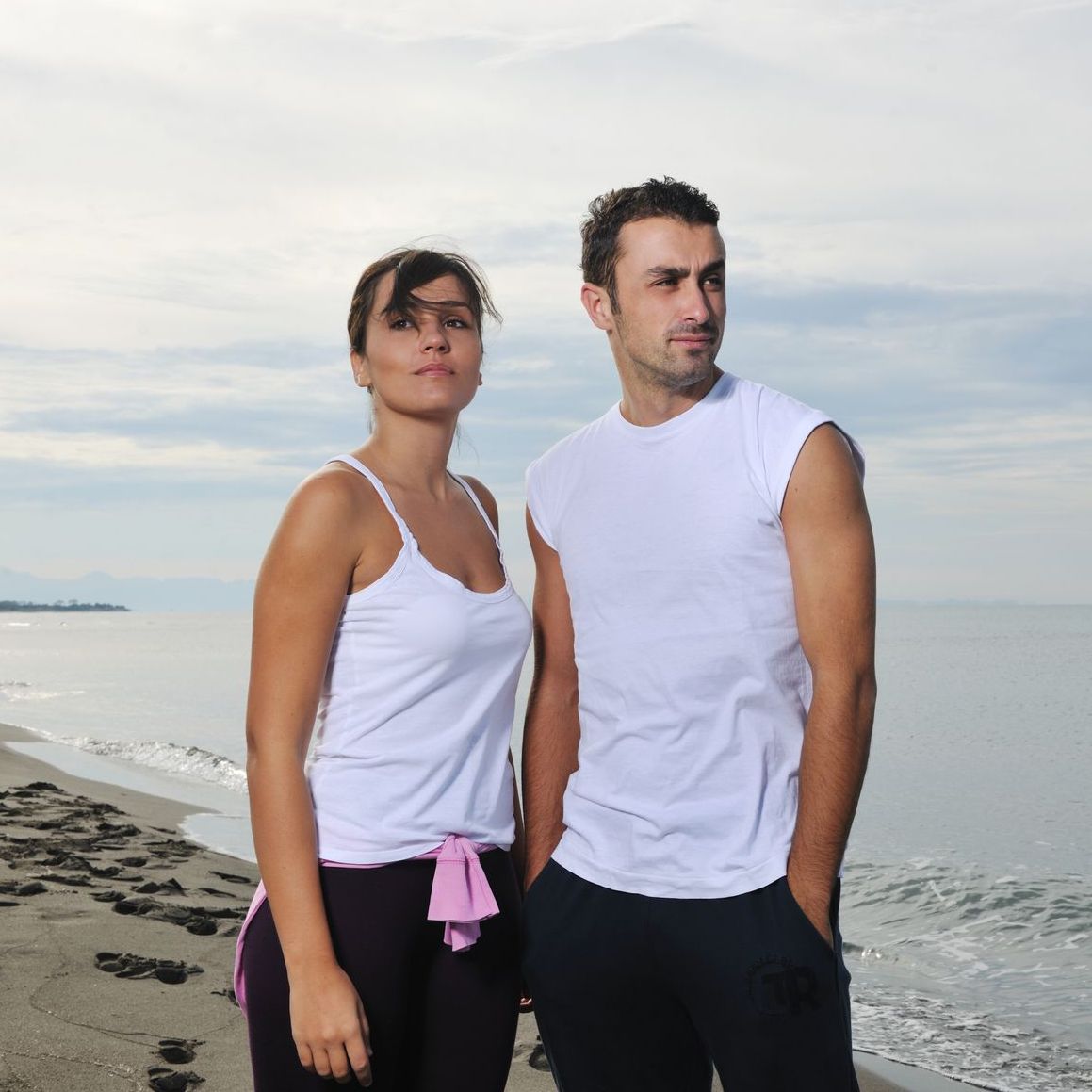 A man and a woman standing on a beach with their hands in their pockets