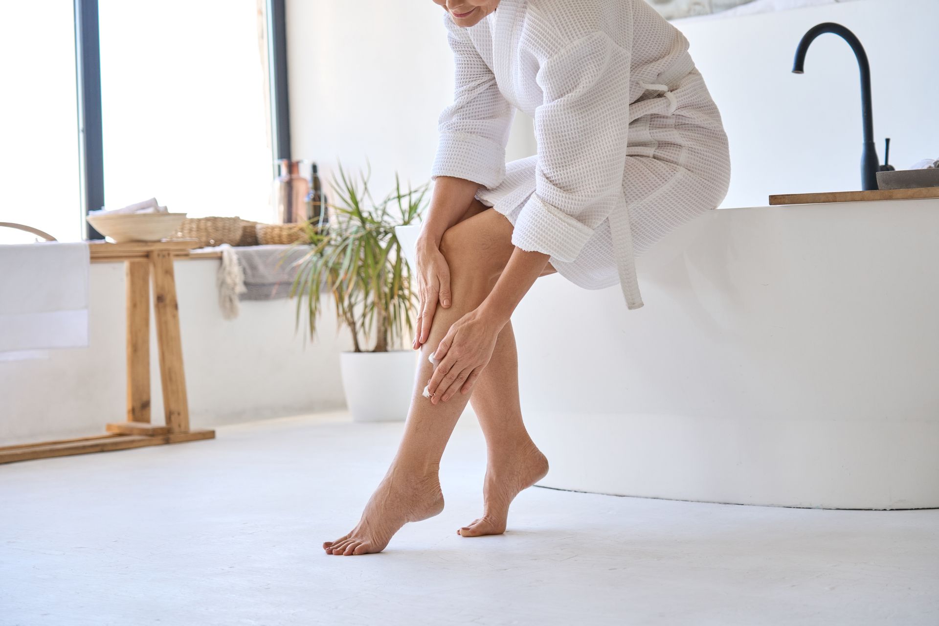 A woman in a bathrobe is sitting on a bathtub and applying lotion to her legs.