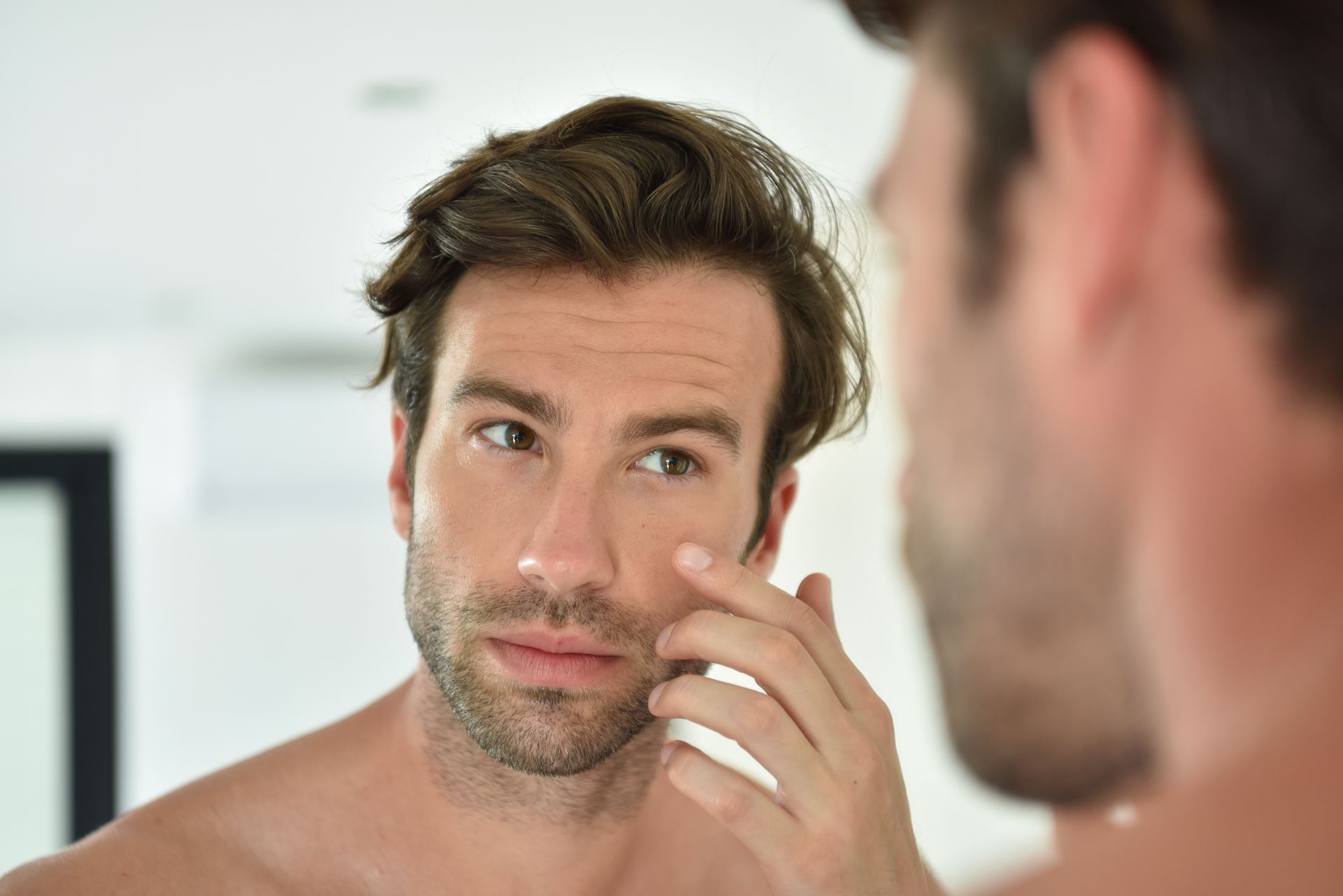 A shirtless man is looking at his face in a mirror.