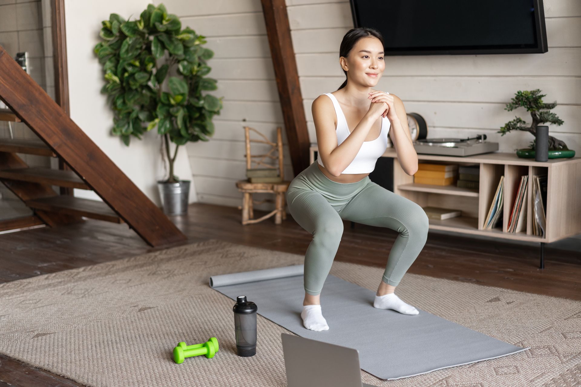 A woman is squatting on a yoga mat in front of a laptop.