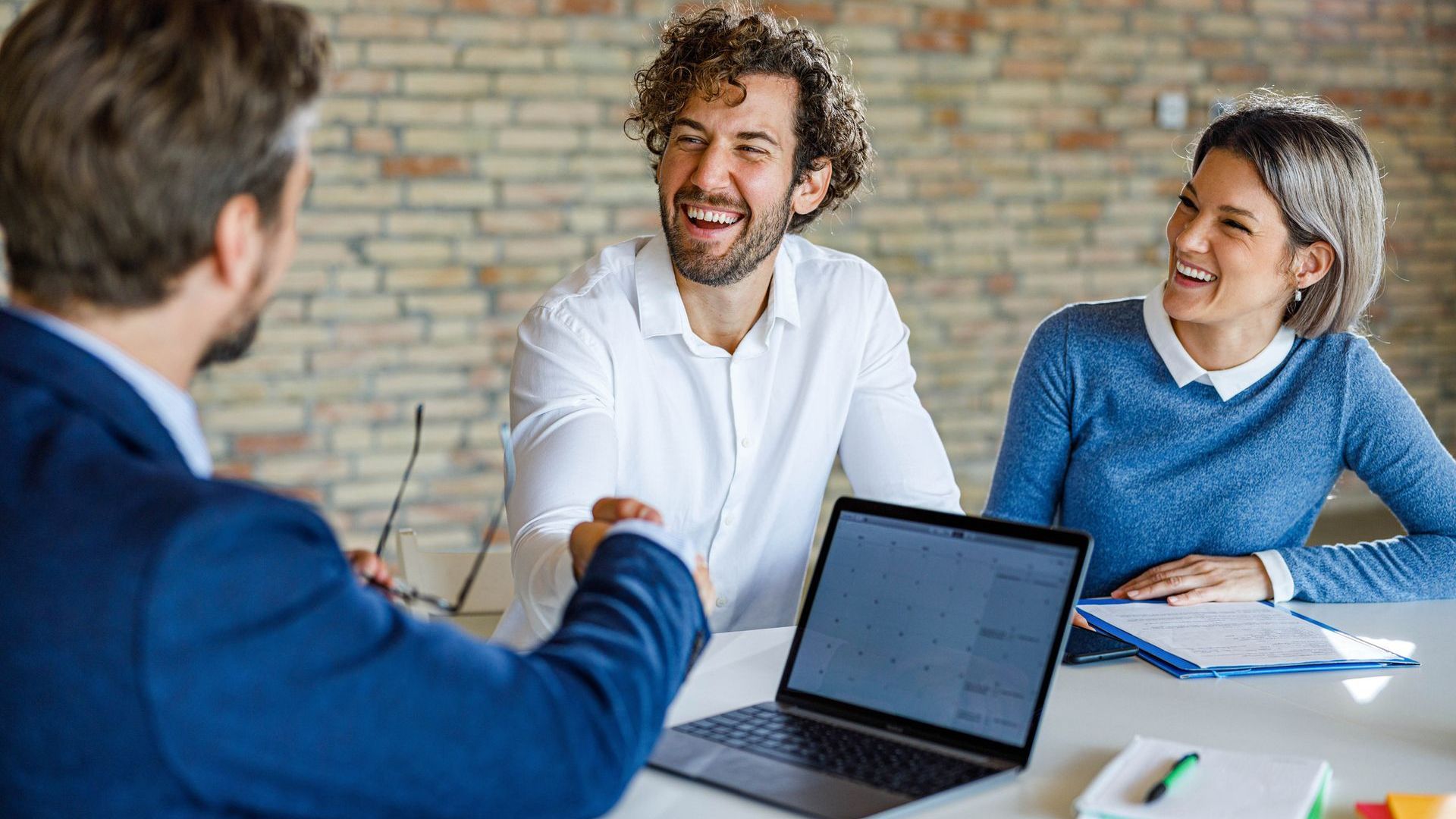 A man is shaking hands with a woman while sitting at a table with a laptop.