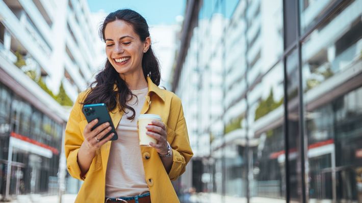 A woman in a yellow jacket is holding a cup of coffee and looking at her phone.