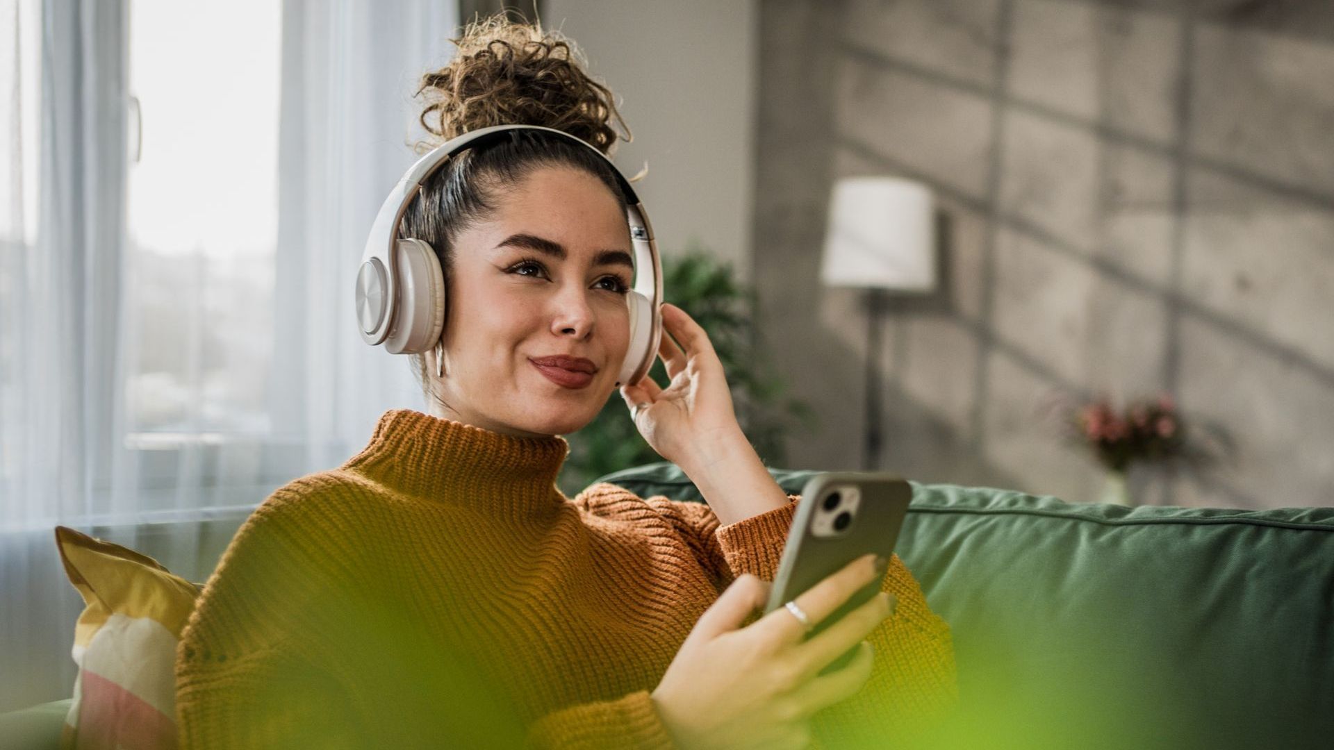 A woman is sitting on a couch wearing headphones and using a cell phone.