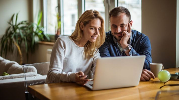 A man and a woman are sitting at a table looking at a laptop computer.