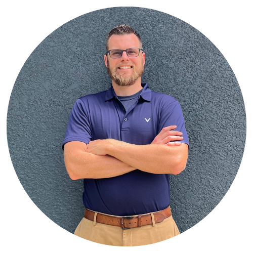 A man wearing glasses and a blue shirt is standing in front of a wall with his arms crossed.