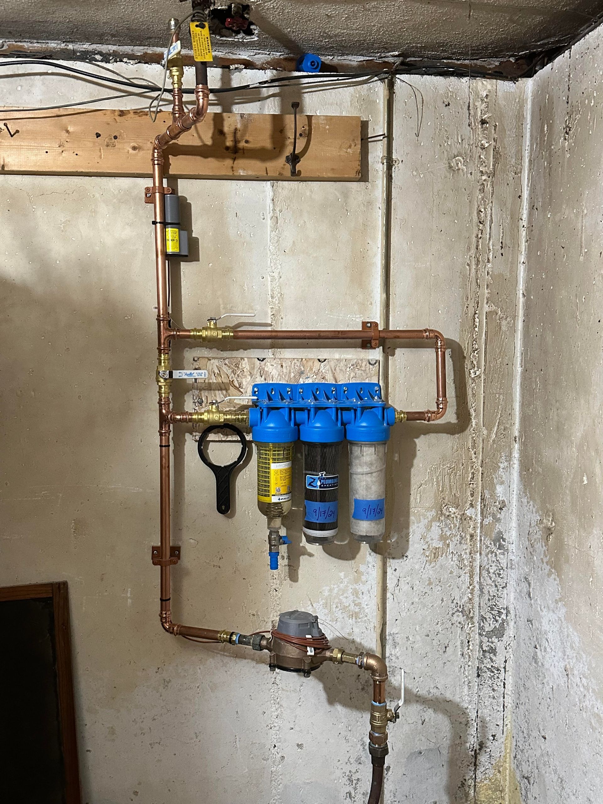 Copper water filtration system attached to a concrete wall, with blue, black, and white filters and a water meter.