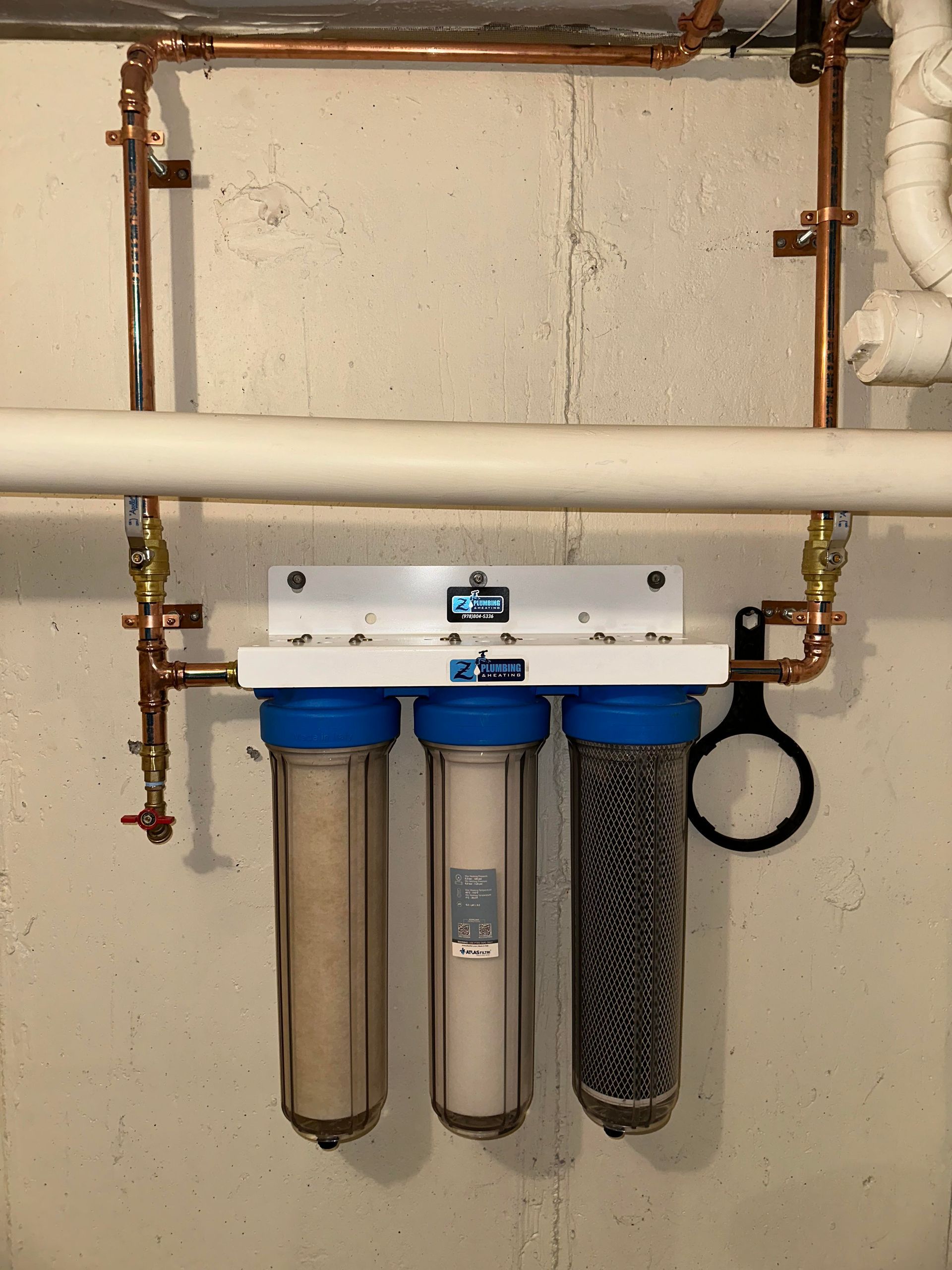 Water filtration system with three clear filter housings, copper piping, and white mounting plate on a concrete wall.