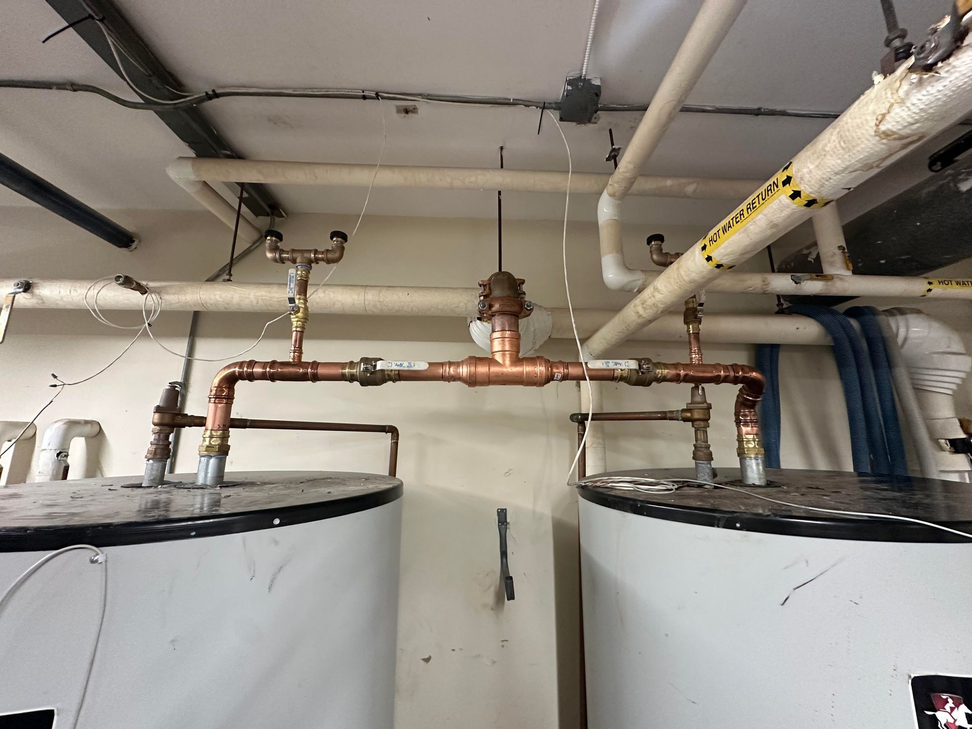 Copper pipes connecting two white water tanks, with a valve and overhead piping in a basement.