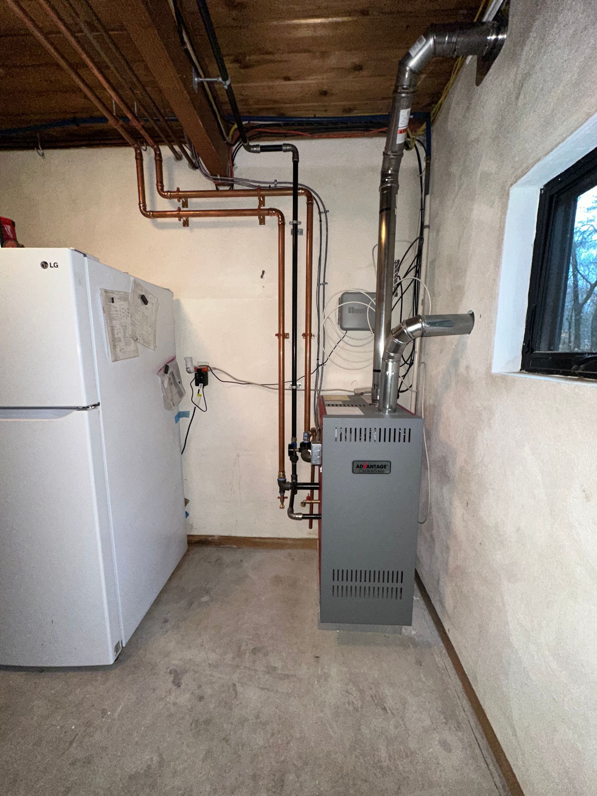 A furnace and refrigerator in a basement, with copper pipes, and a small window.