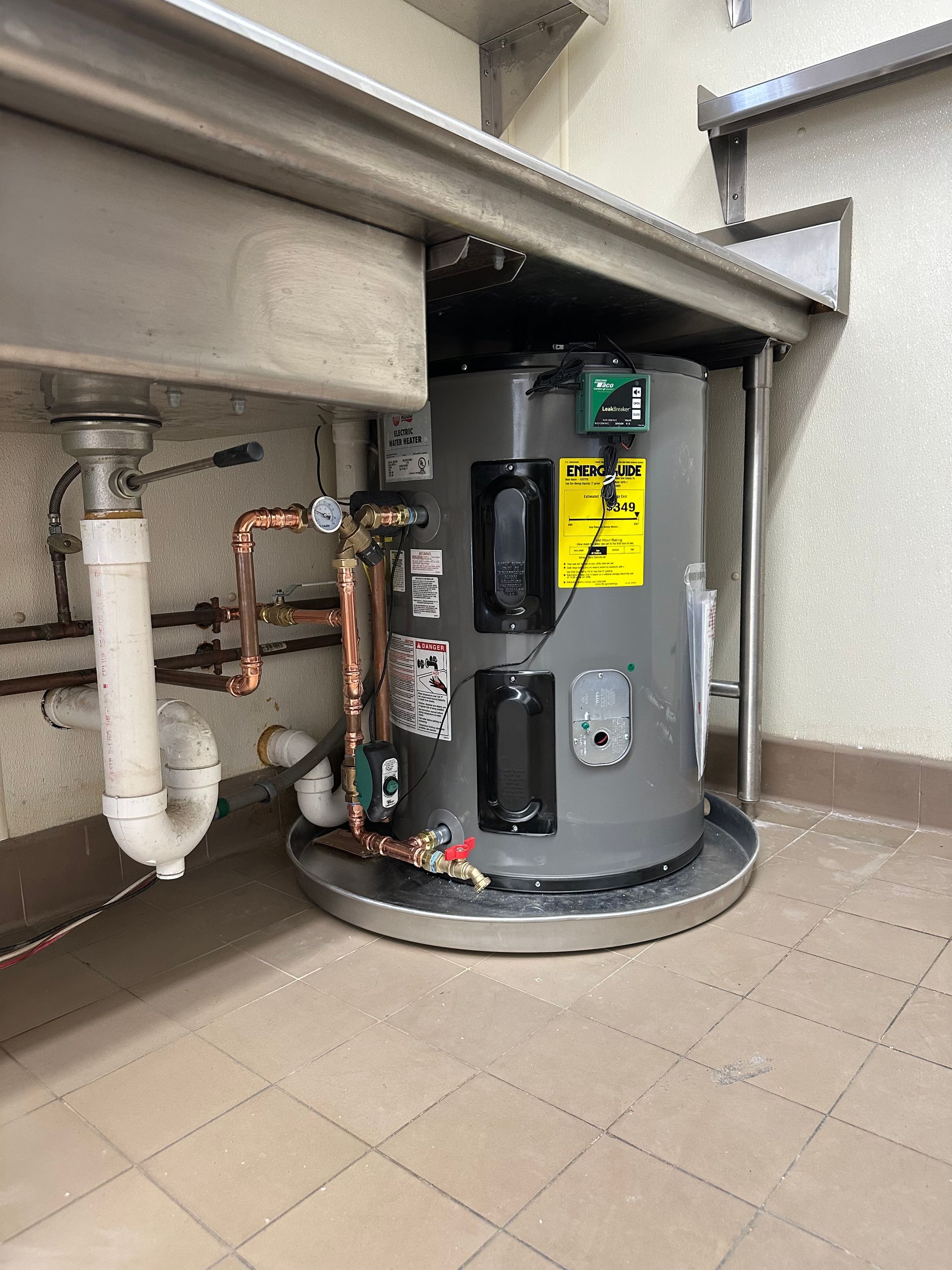 Water heater under a stainless steel sink in a commercial kitchen. Plumbing pipes are visible.