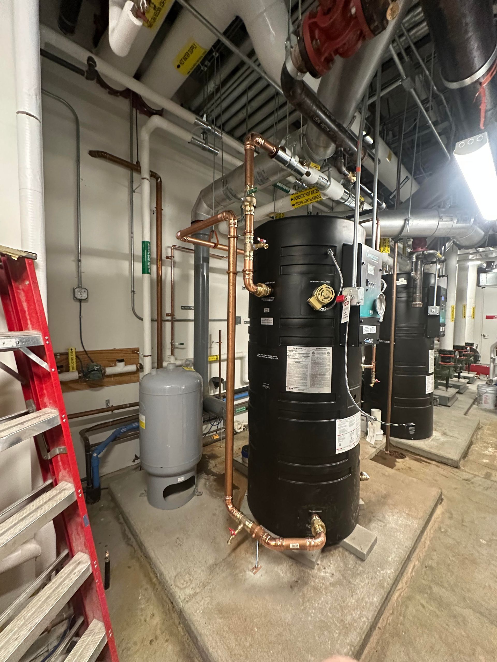 Plumbing equipment in a mechanical room: black tanks, copper pipes, gray expansion tank, red ladder, and white pipes.