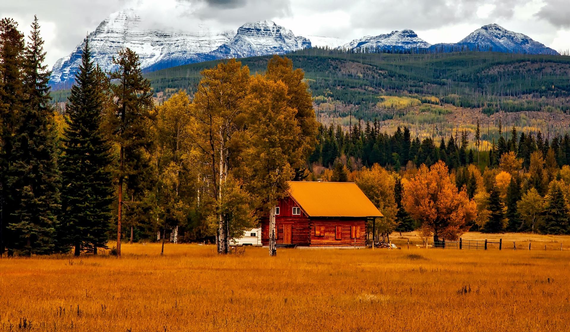 A cabin with an orange roof nestled in a field of dry grass, with a mountain range in the background. Autumn trees surround the cabin.