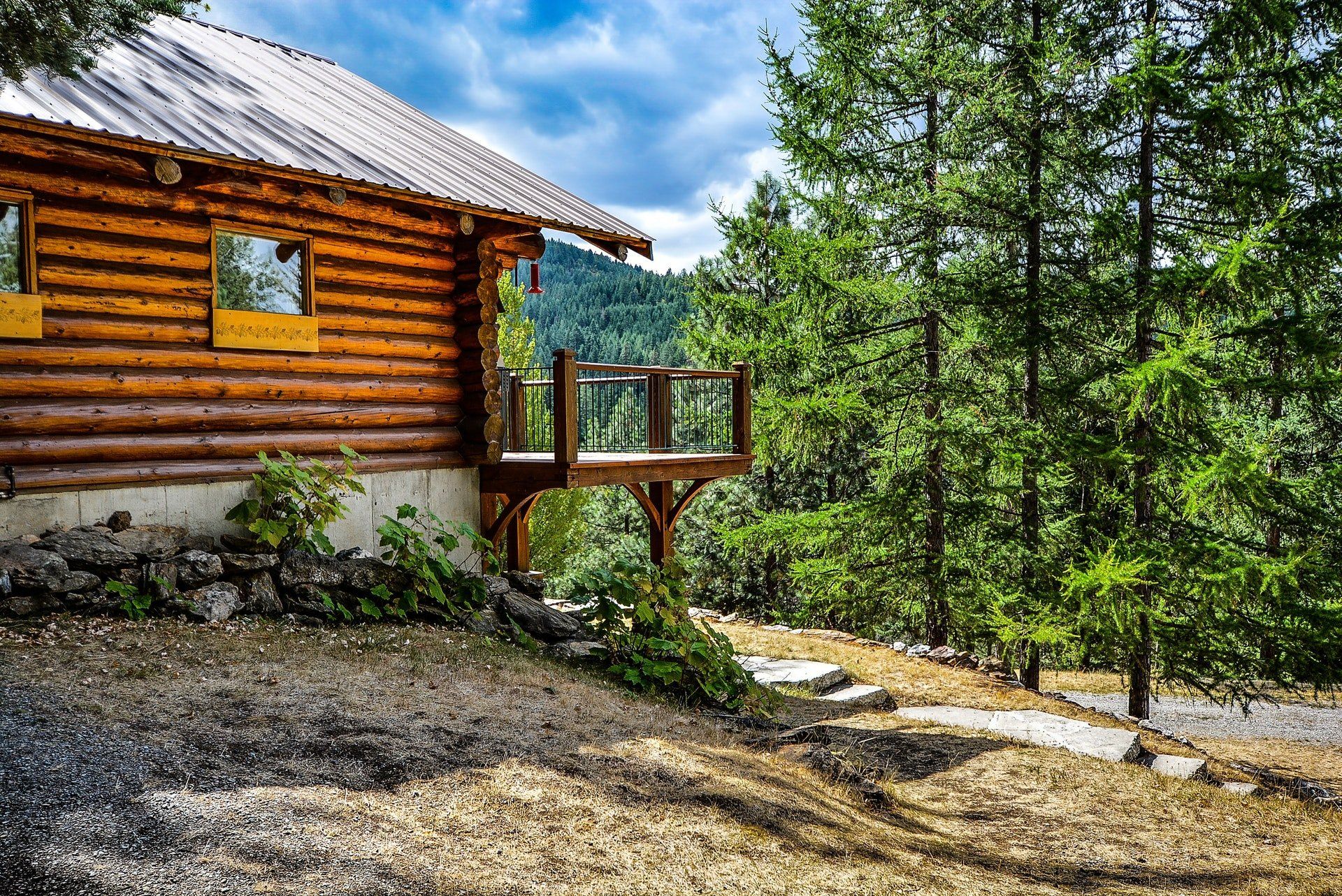 Log cabin with a deck and metal roof surrounded by tall green trees under a cloudy sky.