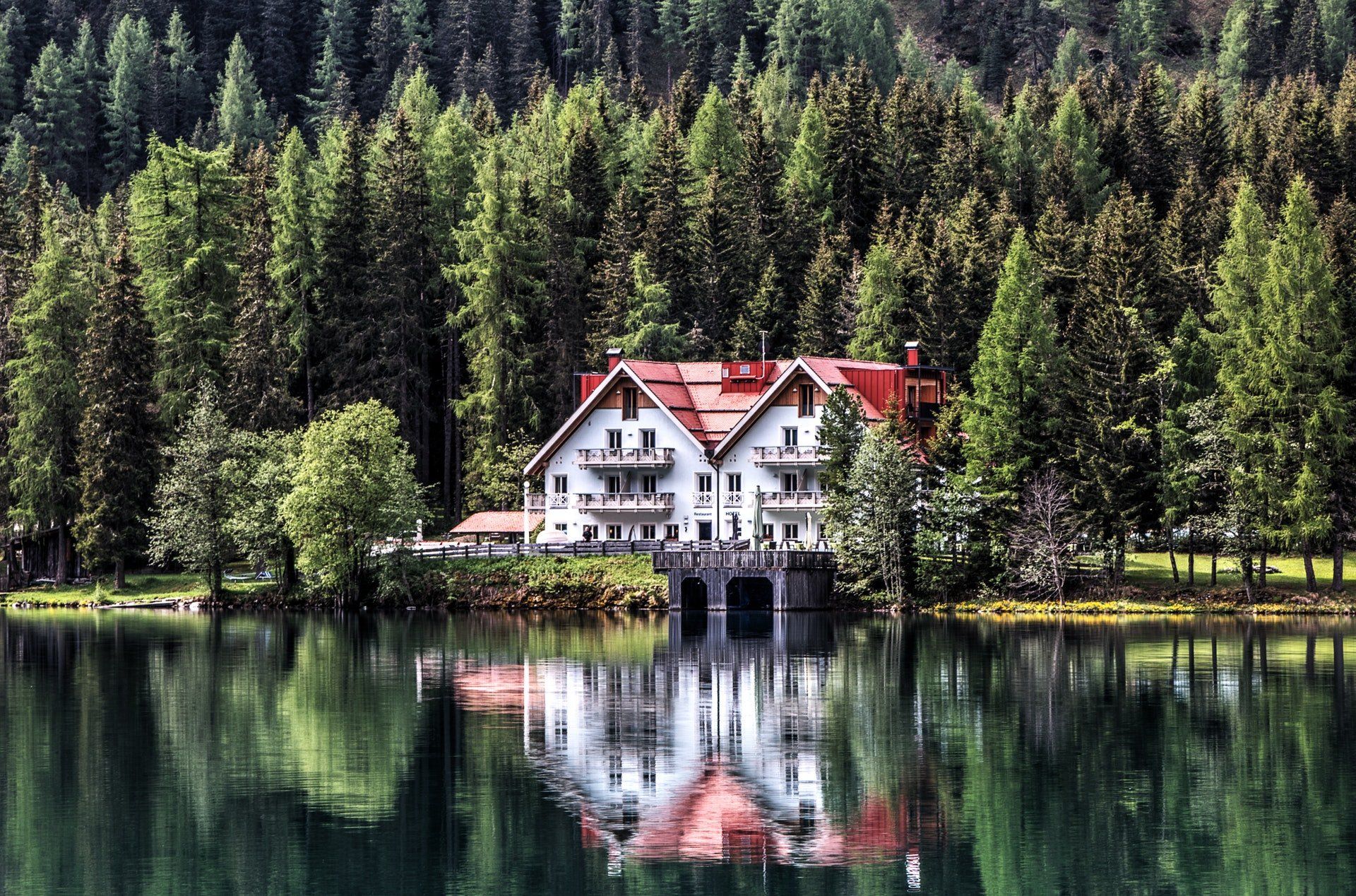 House with white walls and a red roof on a lake, surrounded by a forest of green trees.