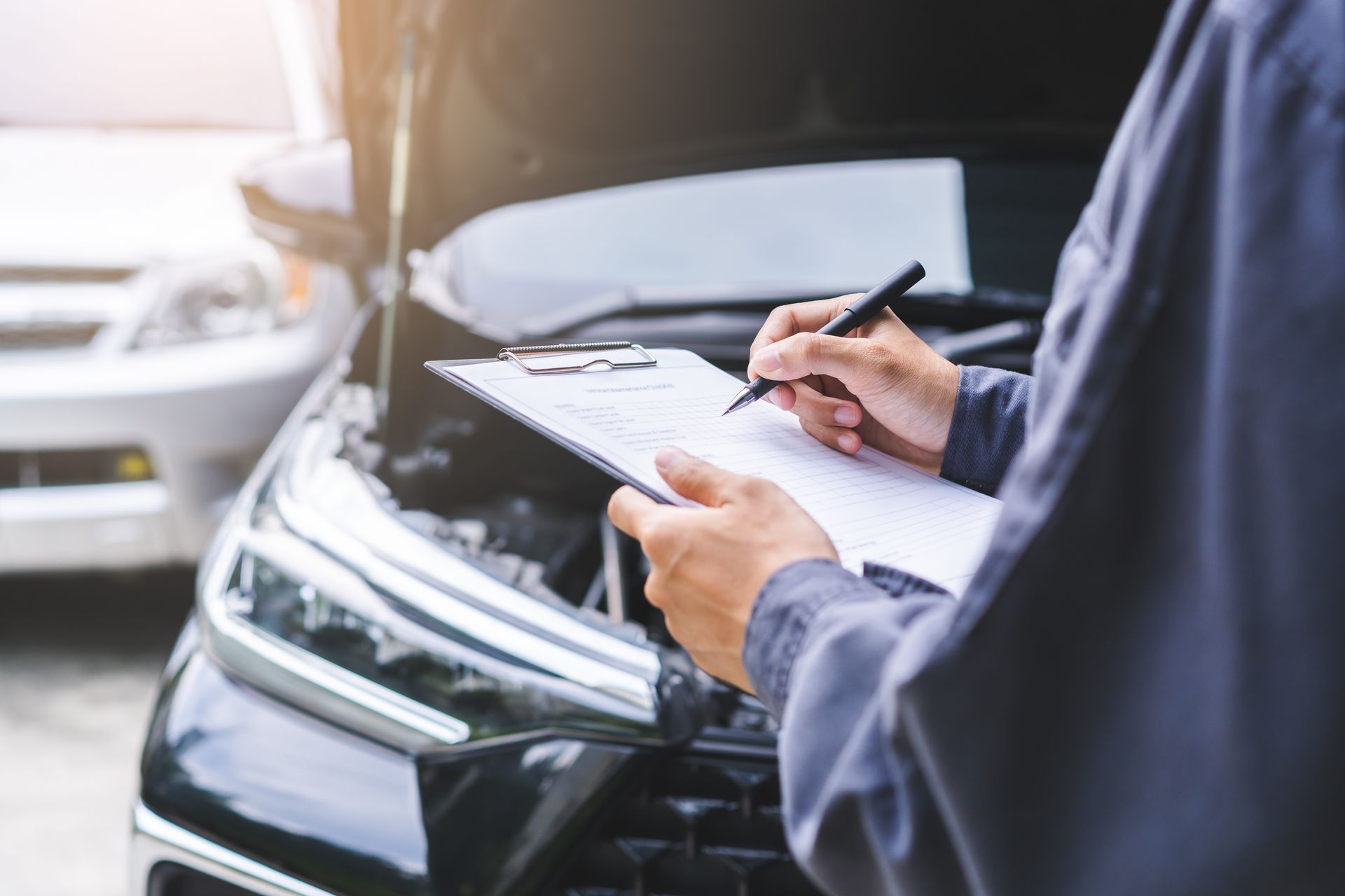 A partial view of a mechanic writing on a clipboard with two cars in the background. A partial view of a mechanic writing on a clipboard with two cars in the background.