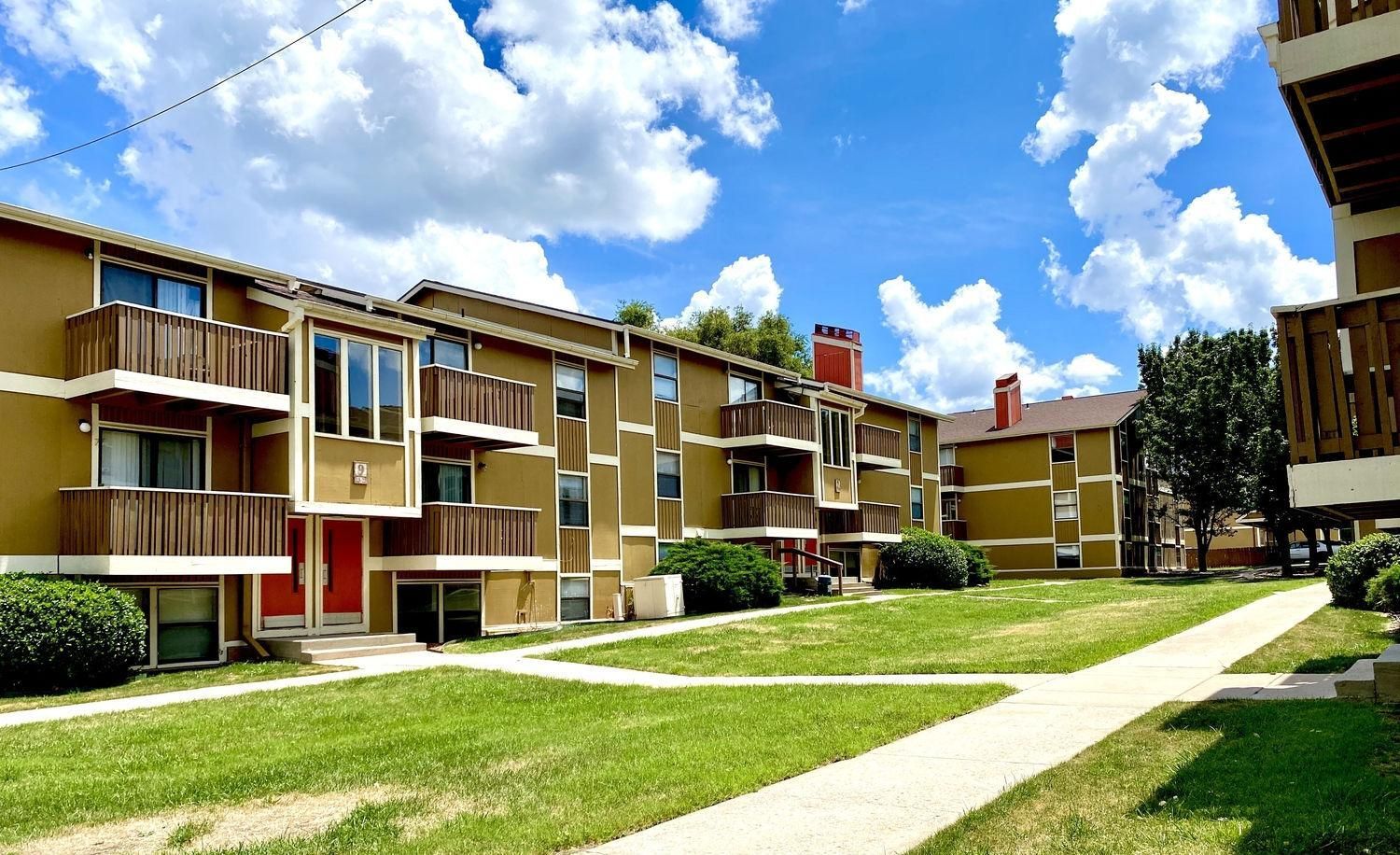 Apartment complex with tan exteriors, wooden balconies, and a green lawn under a bright blue sky with clouds.