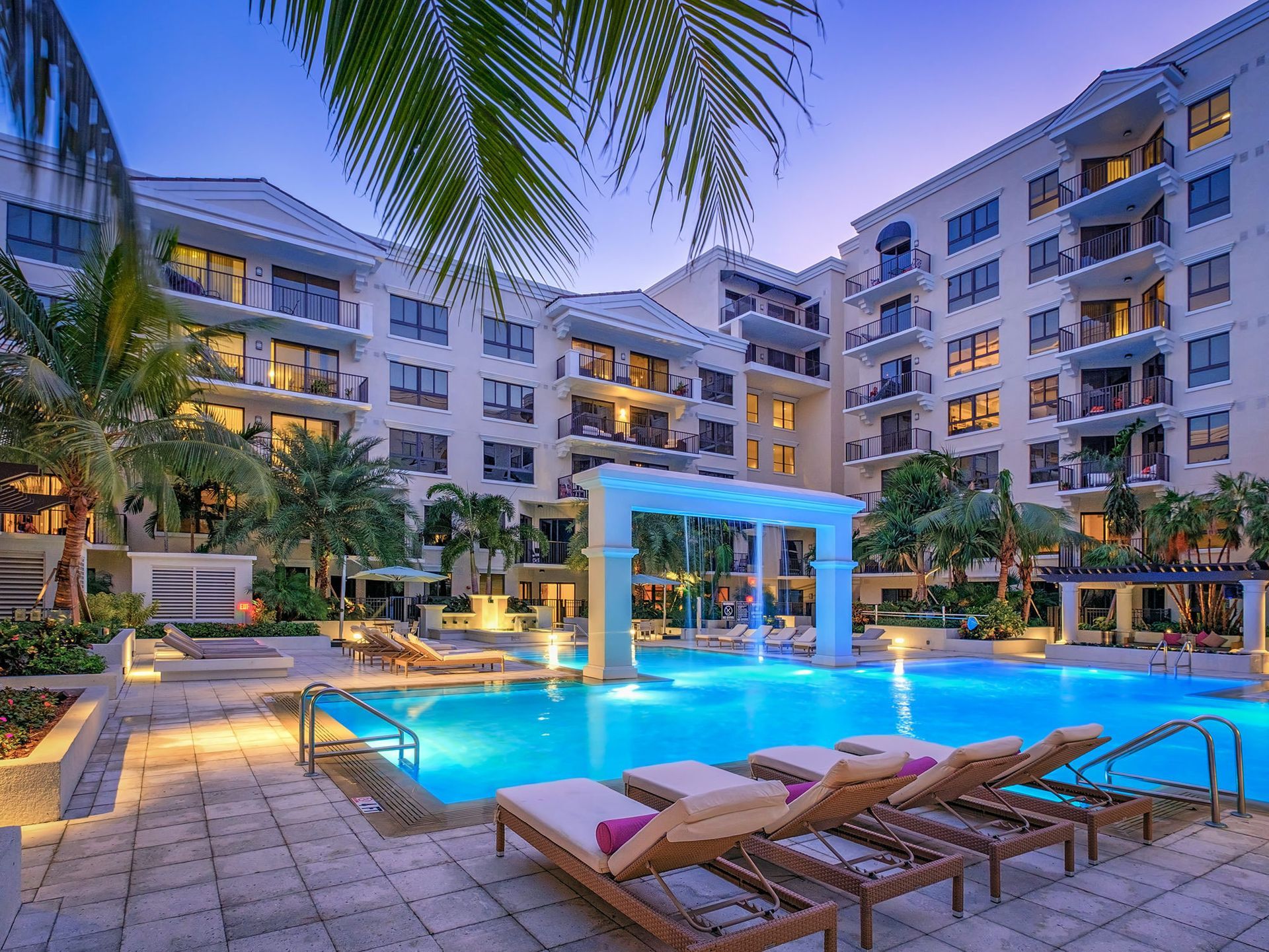 A luxury condo complex with a pool, lounge chairs, and palm trees at dusk. The architecture features white stucco and balconies.