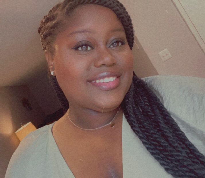 Woman with braided hair smiling, wearing a necklace and a light-colored top.