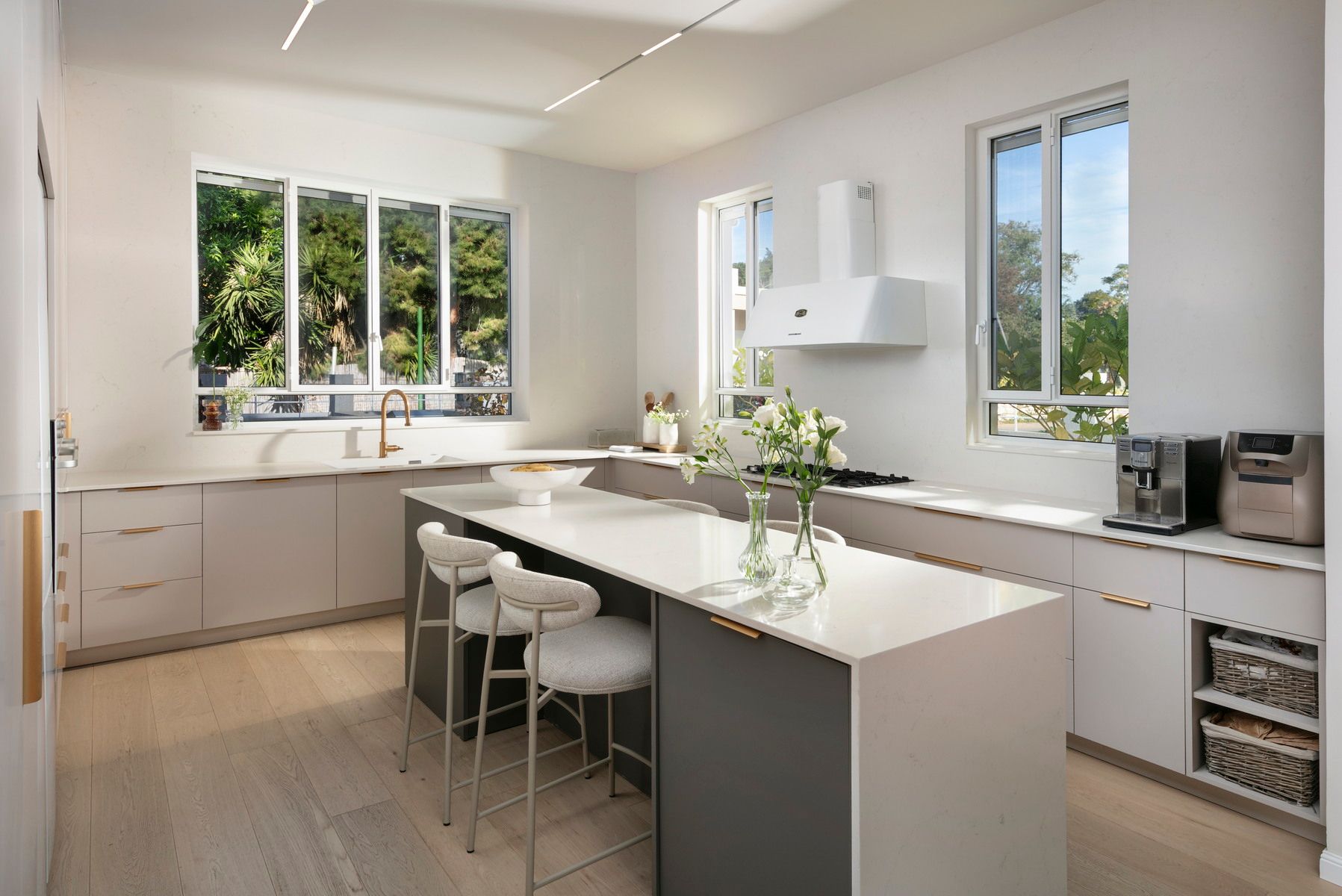 מטבח לבן מודרני עם אי, ארונות אפורים, חלונות גדולים וכסאות בר.