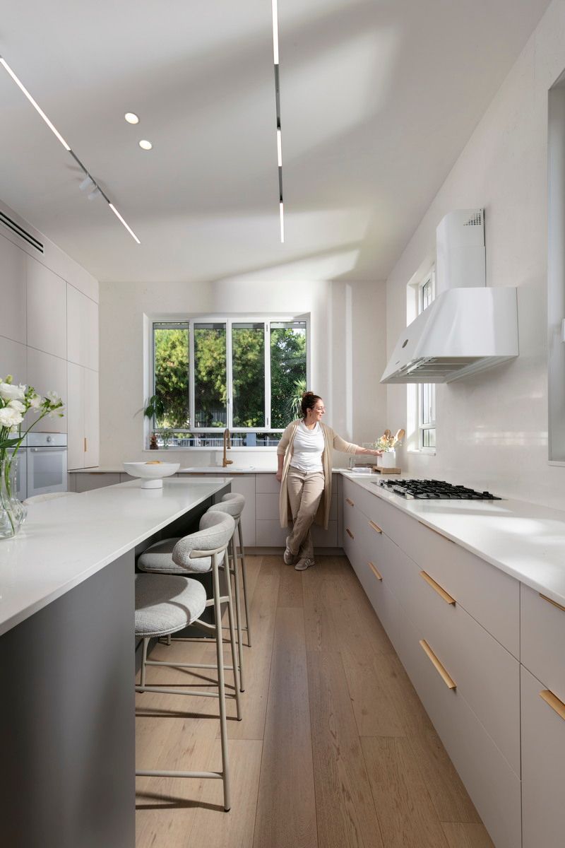 מטבח מודרני עם אישה שנוגעת במשטח העבודה. ארונות בהירים, אי עם שרפרפים וחלון גדול.