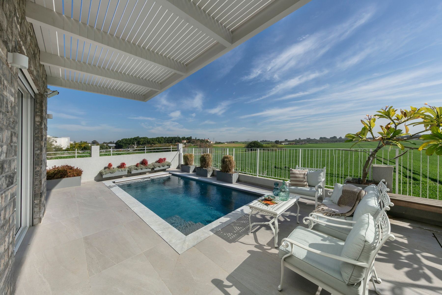 אזור בריכה ופטיו עם מקומות ישיבה, שולחן קטן וצמחייה ירוקה המשקיפה על נוף מרוחק תחת שמיים כחולים.