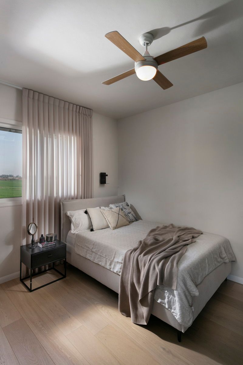 חדר שינה עם מיטה, קירות בצבעים בהירים, ריצוף דמוי עץ, מאוורר תקרה וחלון עם וילונות בצבע בז'.