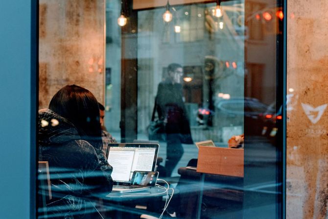 Person working on a laptop inside a cafe, reflected in the window, with a pedestrian walking by.