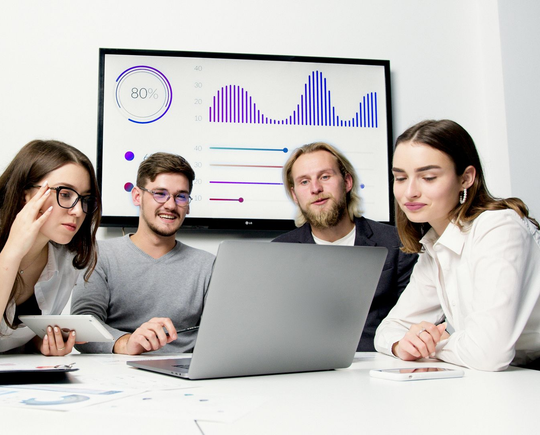 Four people at a table reviewing data on a laptop and a large screen, in a modern office.