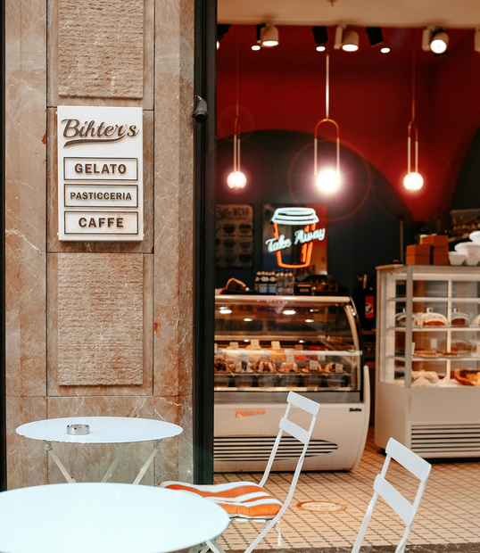 A gelato shop entrance: white tables and chairs outside, display case with treats, red interior.